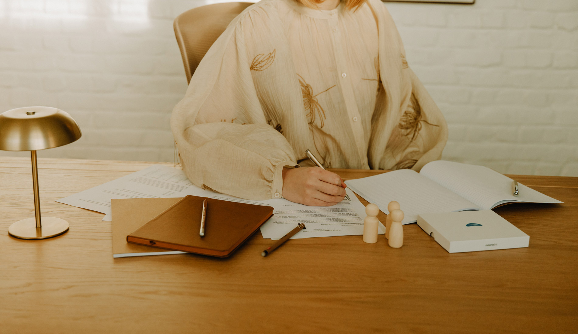 Persoon in beige blouse schrijft met een pen op documenten aan een houten bureau met notitieboekjes, pennen, houten poppetjes en een gouden bureaulamp.