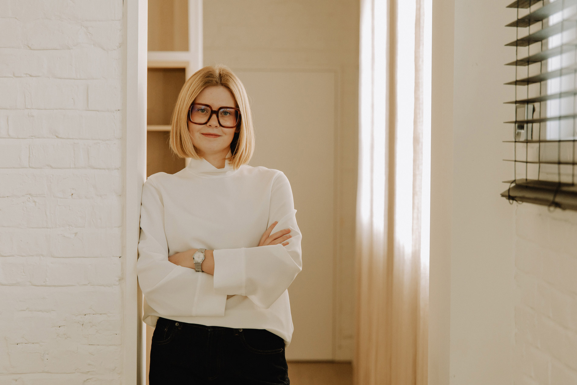 Vrouw met blond haar en grote bril, gekleed in een witte trui, leunt met gekruiste armen tegen een witte muur in een lichte kamer.