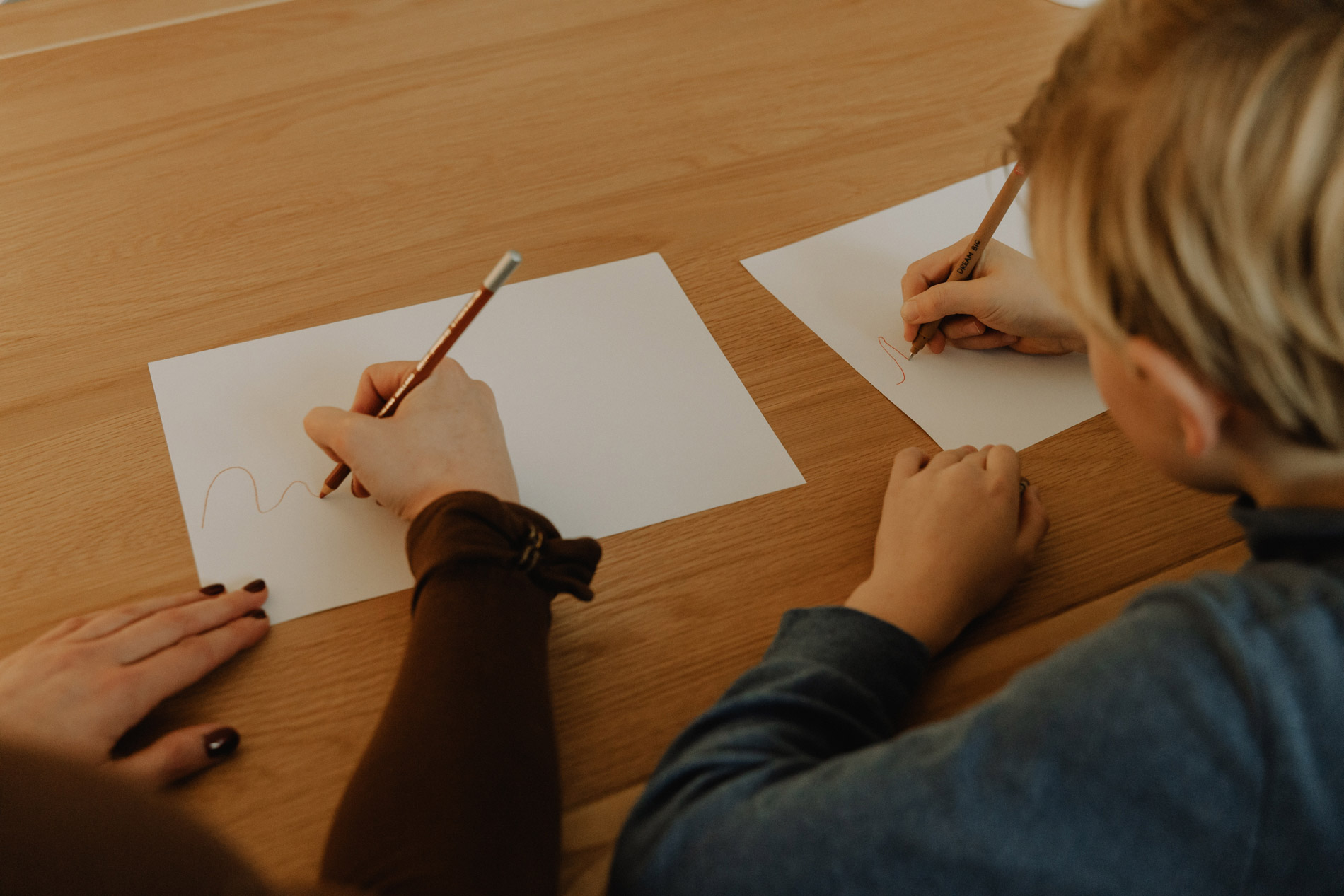 Een volwassene en een kind tekenen met potlood op blanco papier op een houten tafel.