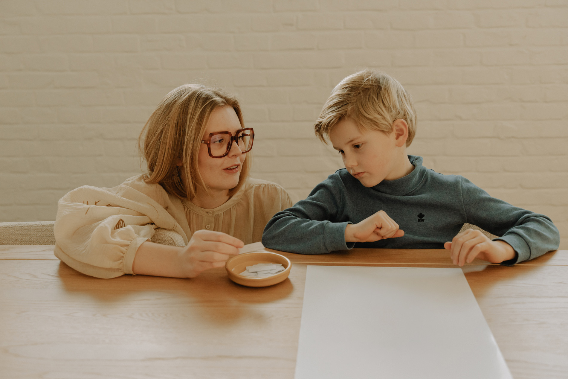 Een vrouw met grote bril en een jongen zitten samen aan een houten tafel met een tekening van een vuurtoren en een kom met papiertjes.
