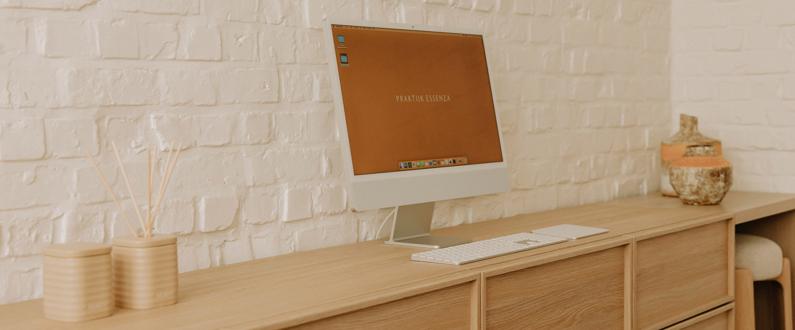 Moderne computer met het scherm 'Praktijk Essenza' op een houten bureau tegen een witte bakstenen muur, met geurstaafjes en keramische vazen.