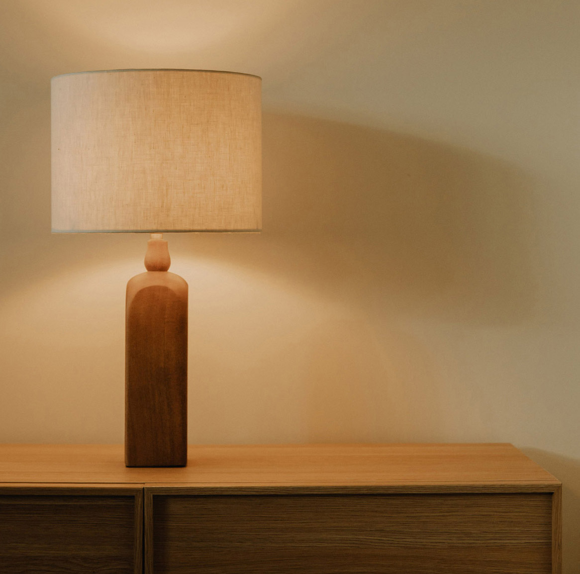 Een houten tafellamp met een beige lampenkap staat op een houten kast tegen een effen beige muur.