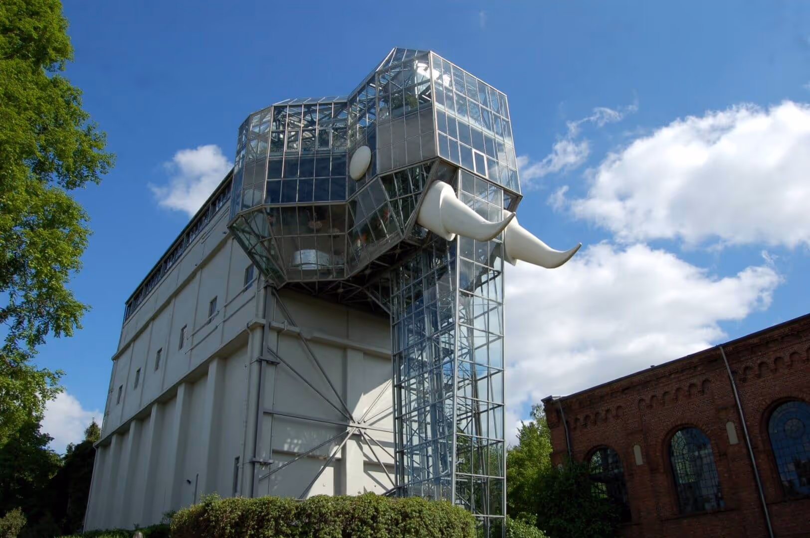 Gebäude mit einer großen gläsernen Struktur, die einem Elefantenkopf mit Stoßzähnen ähnelt, bei klarem Himmel.