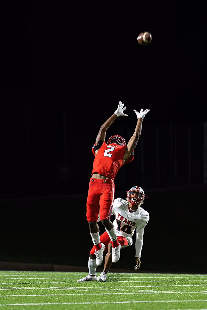Football player in red jersey number 2 leaping to catch a football with an opposing player in a white jersey number 14 behind him on a lit field at night.