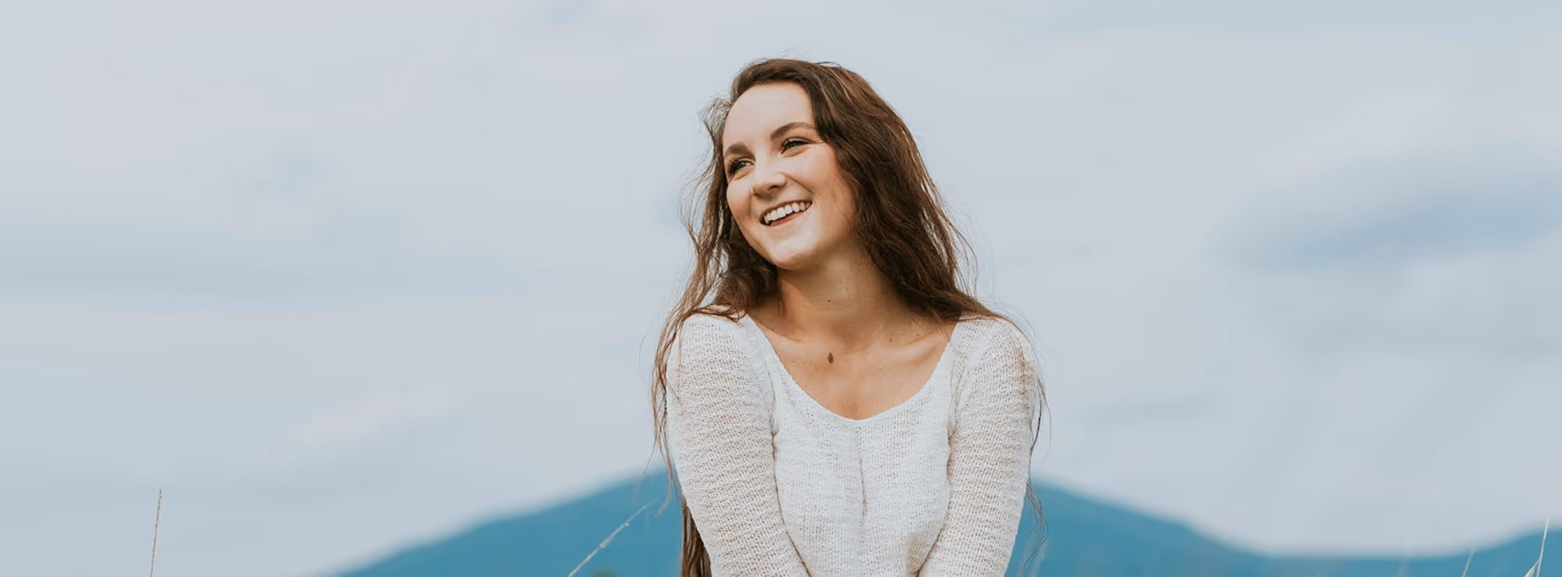 Smiling young woman with long brown hair wearing a white sweater outdoors with mountains and sky in the background.