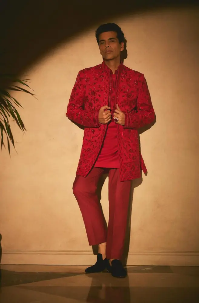 Filmmaker Karan Johar wearing a custom crimson red Manish Malhotra sherwani with intricate floral threadwork and tonal embroidery, paired with matching slim-fit trousers.