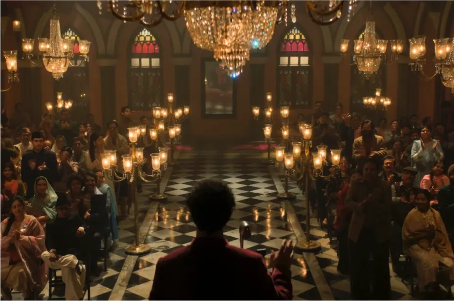 A grand Mushaira (poetry session) scene from the film Gustaakh Ishq, featuring a poet performing on stage before a large audience in a lavishly lit hall with chandeliers.