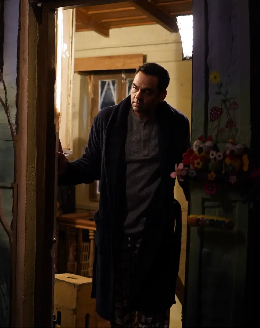 Actor Abhay Deol playing Sidhant, standing in a rustic wooden doorway next to a colorful "Welcome" sign during a scene in Bun Tikki.