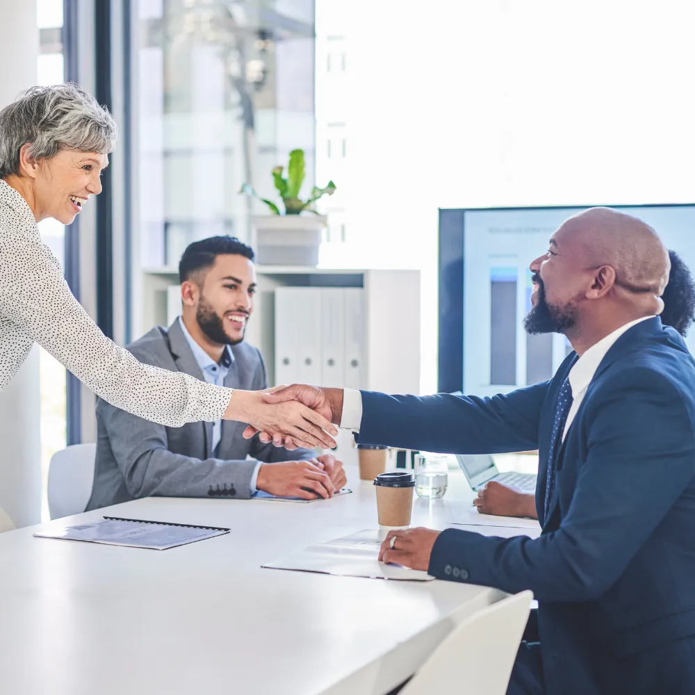 Two professionals shaking hands at a meeting table while another person observes.