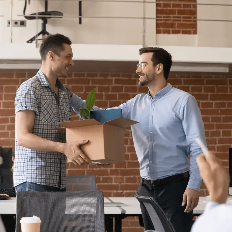 Two men smiling and shaking hands in an office, one holding a box with personal items.