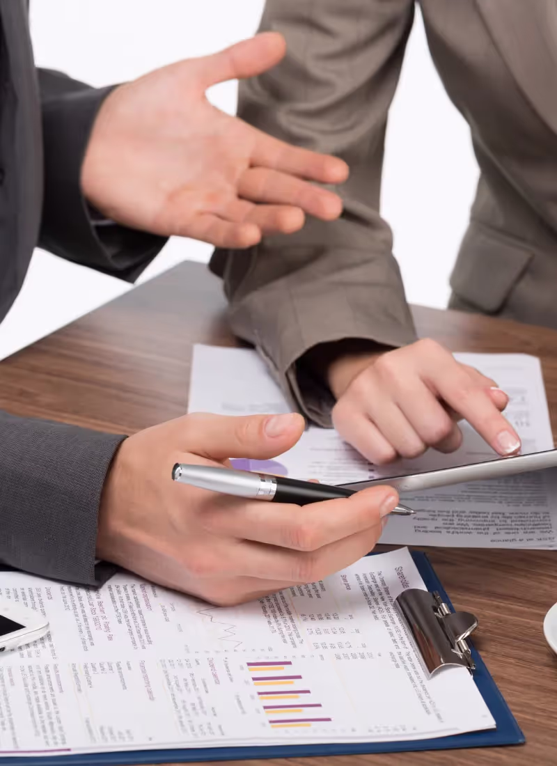 Two professionals discussing over a document with graphs, one gesturing while the other holds a pen and points at the paper.