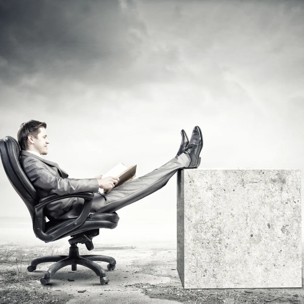 Businessman reading a book with feet propped up on a concrete block, outdoors in a desolate landscape.