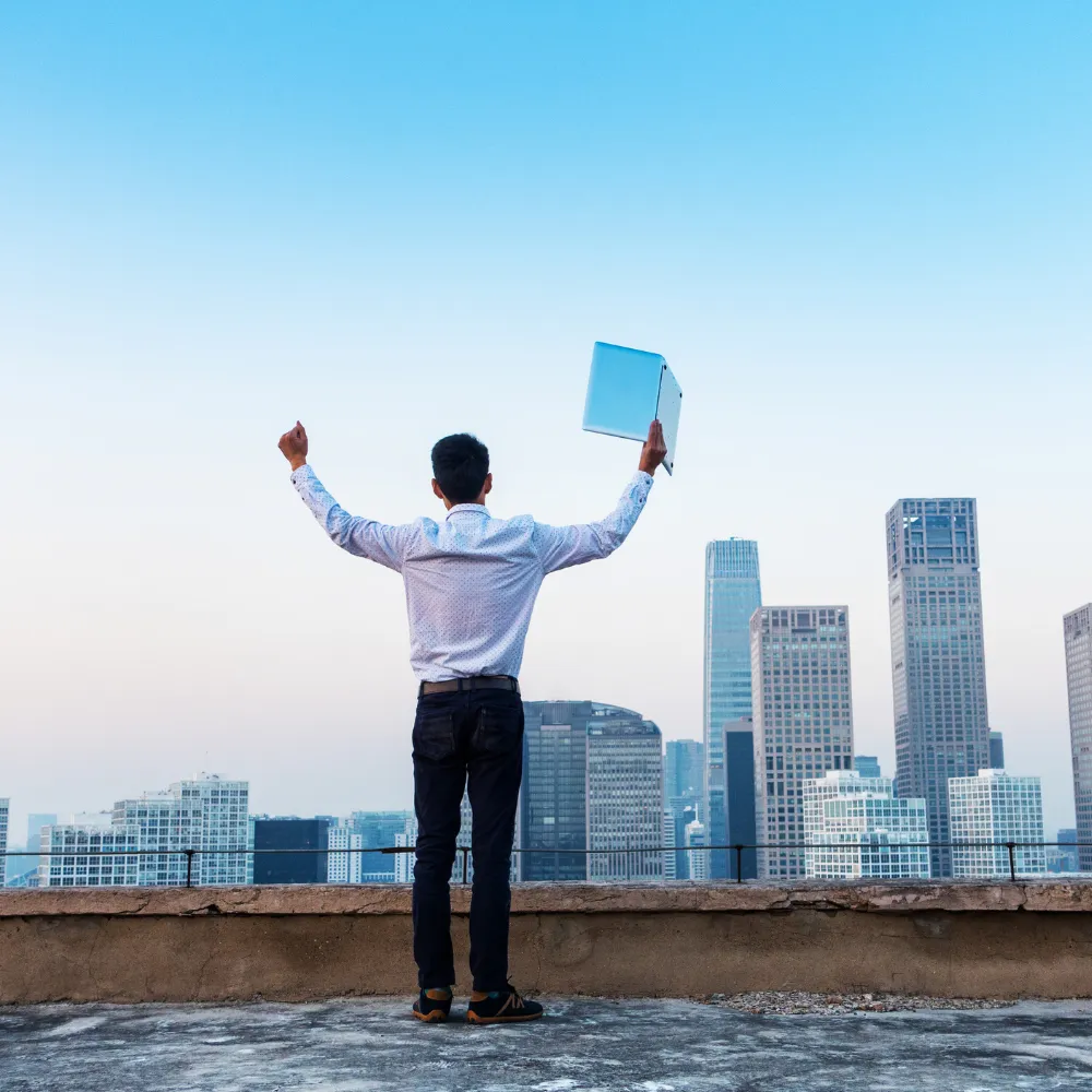 Man standing on a rooftop with arms raised and holding a document, overlooking a city skyline at dusk.