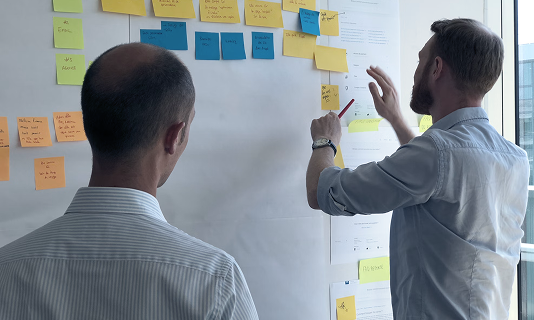 Two men collaborating in an office, discussing colorful sticky notes placed on a whiteboard.
