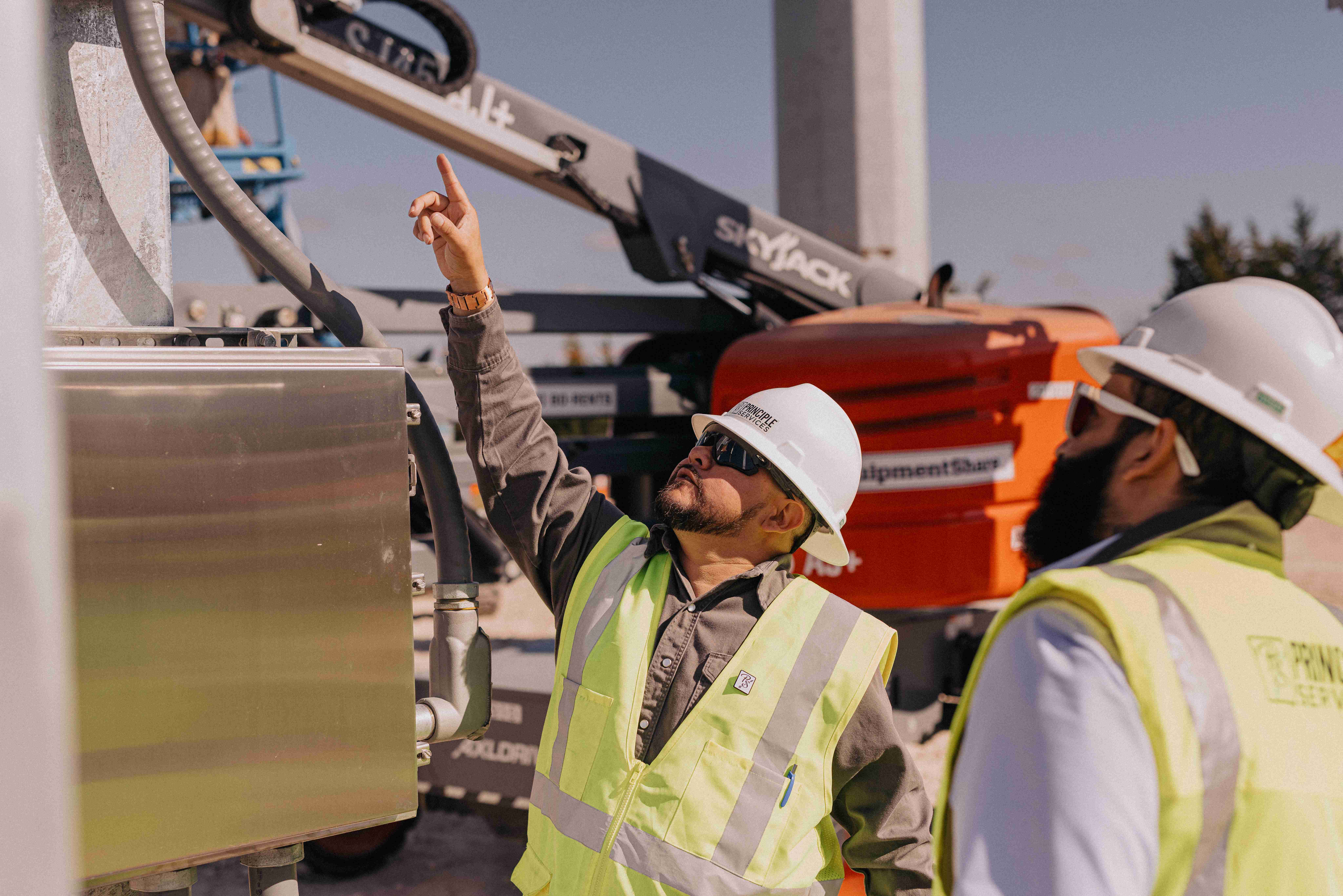Three construction workers wearing safety helmets and high-visibility vests, standing at an electrical substation site.