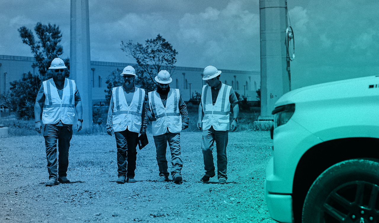 Five construction workers wearing hard hats and reflective safety vests standing in front of electrical infrastructure.
