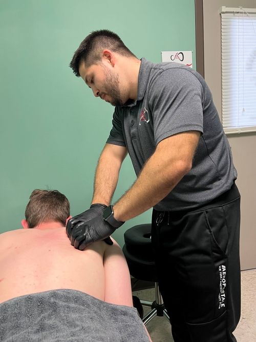 Therapist wearing black gloves performing a back massage on a shirtless person lying face down under a gray towel in a treatment room.