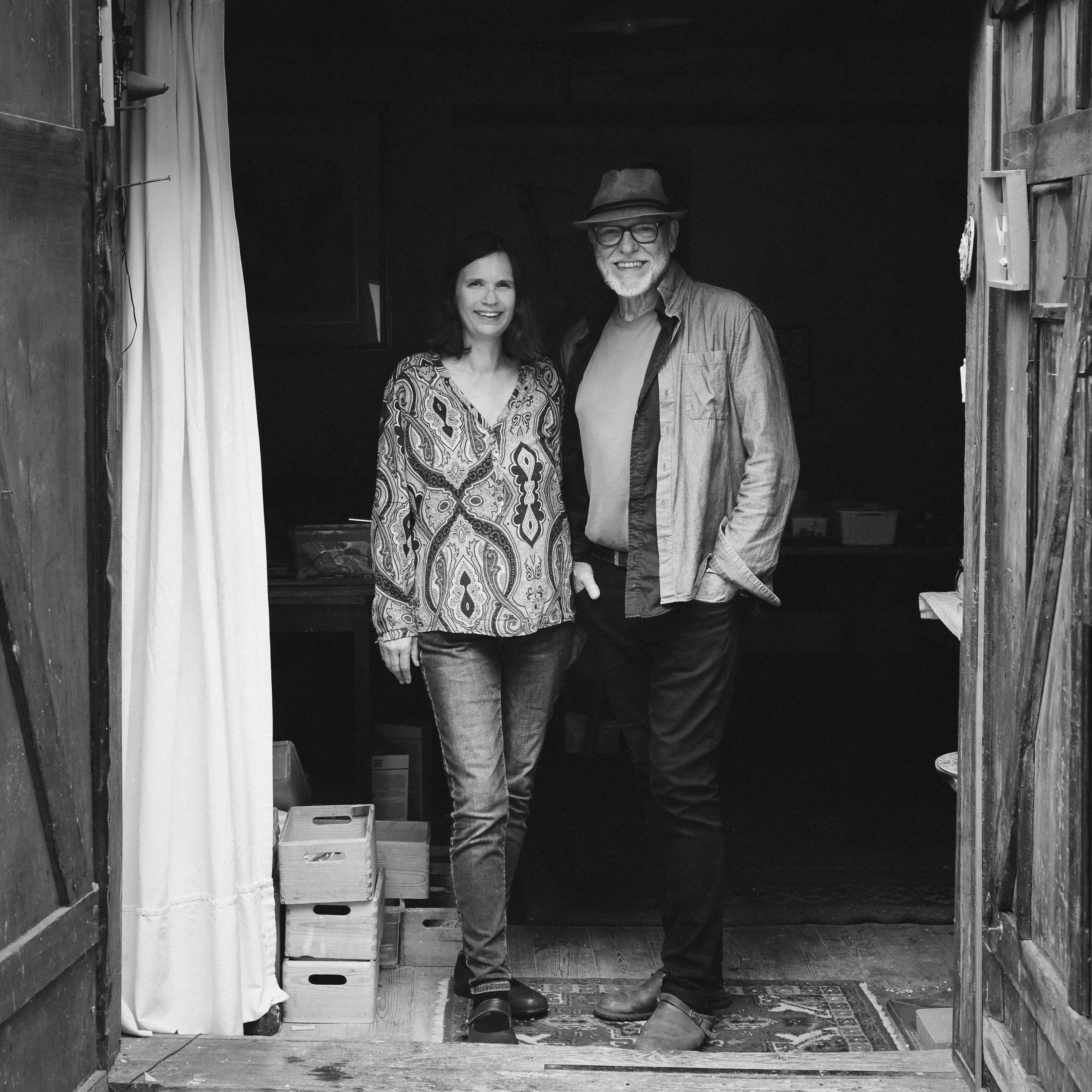 Smiling middle-aged man wearing a hat and glasses stands next to a smiling woman in patterned blouse at an open wooden doorway.