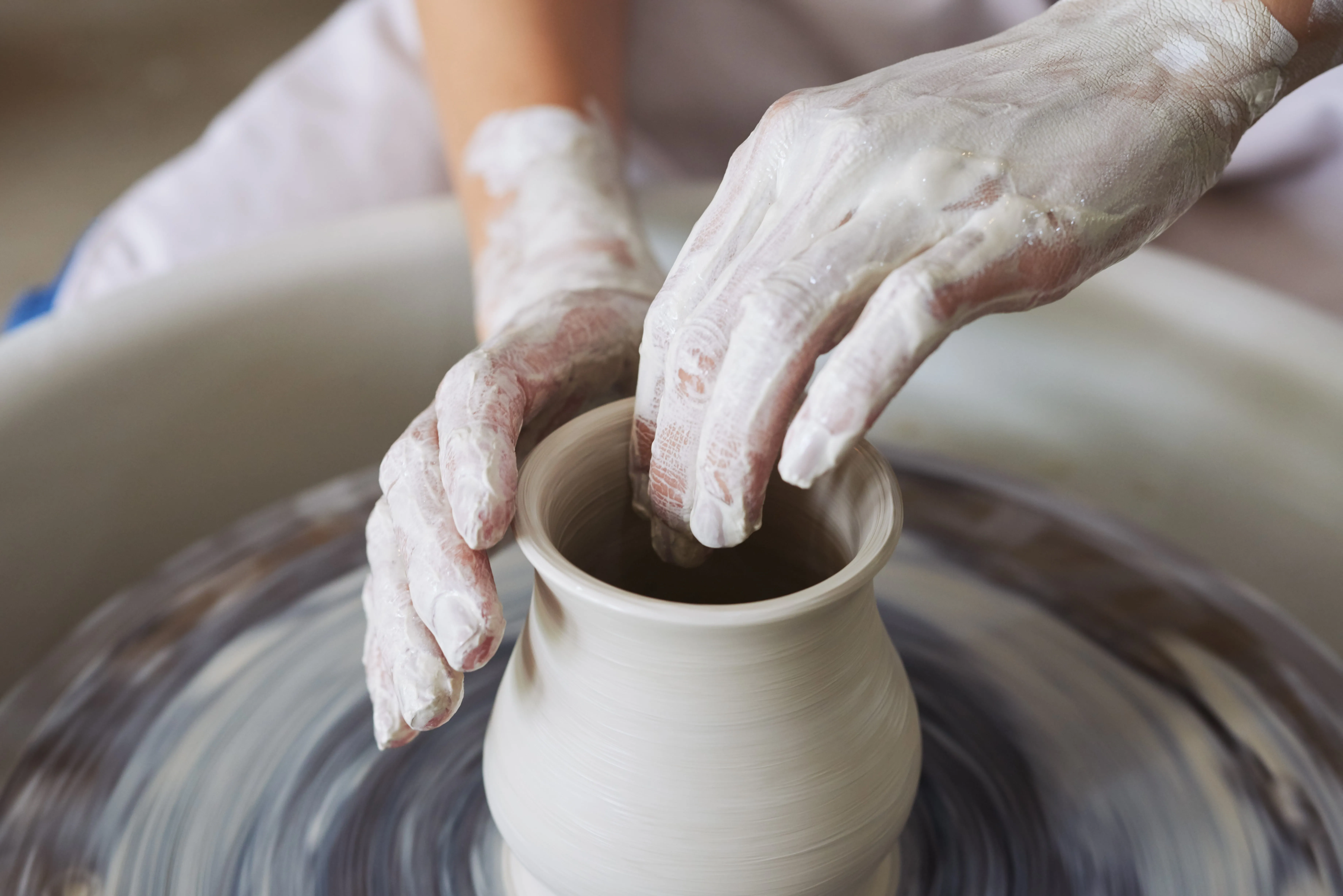Hands covered in clay shaping a pot on a spinning pottery wheel.