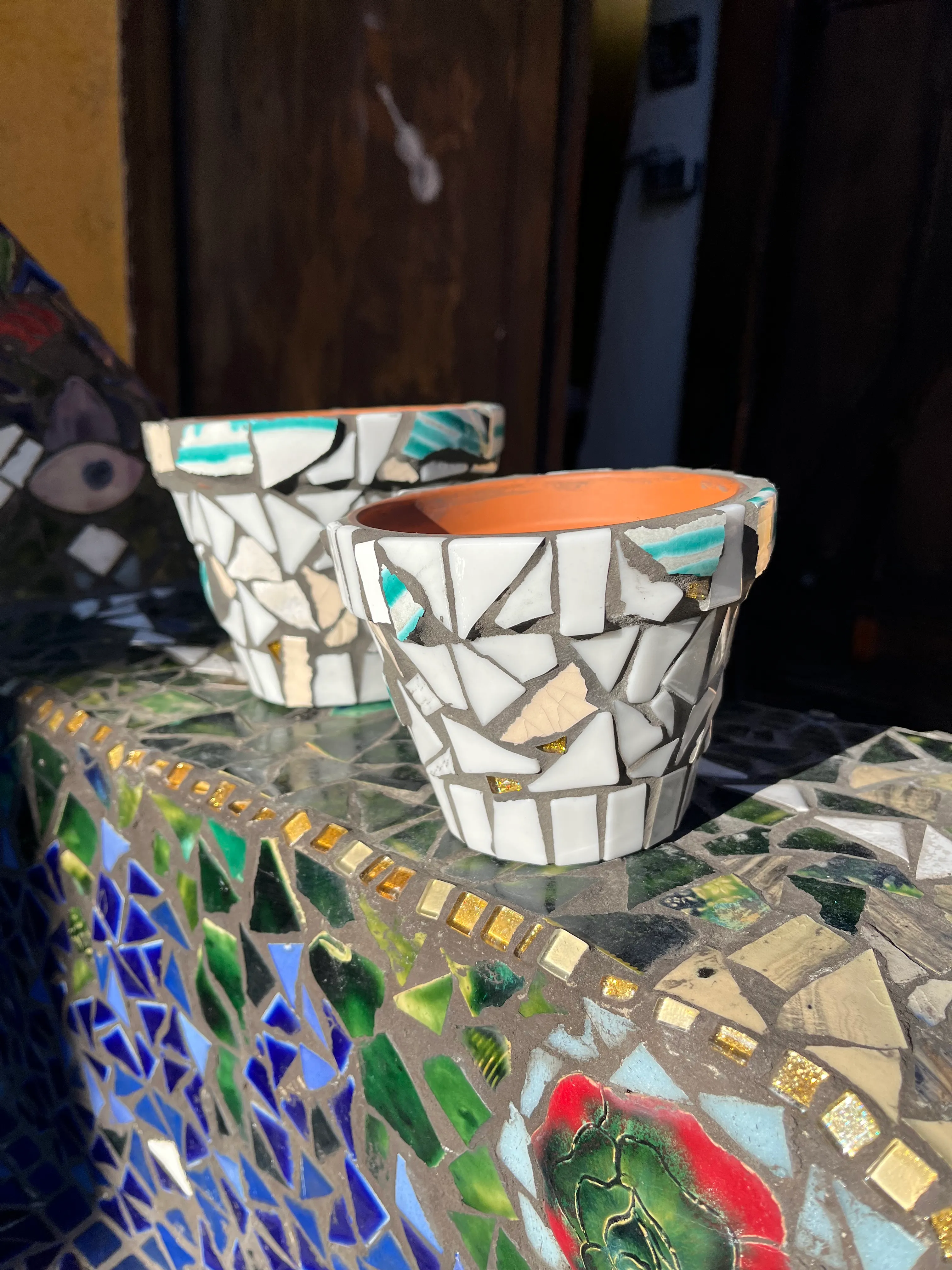 Two mosaic-patterned flower pots on a colorful mosaic table bathed in sunlight.
