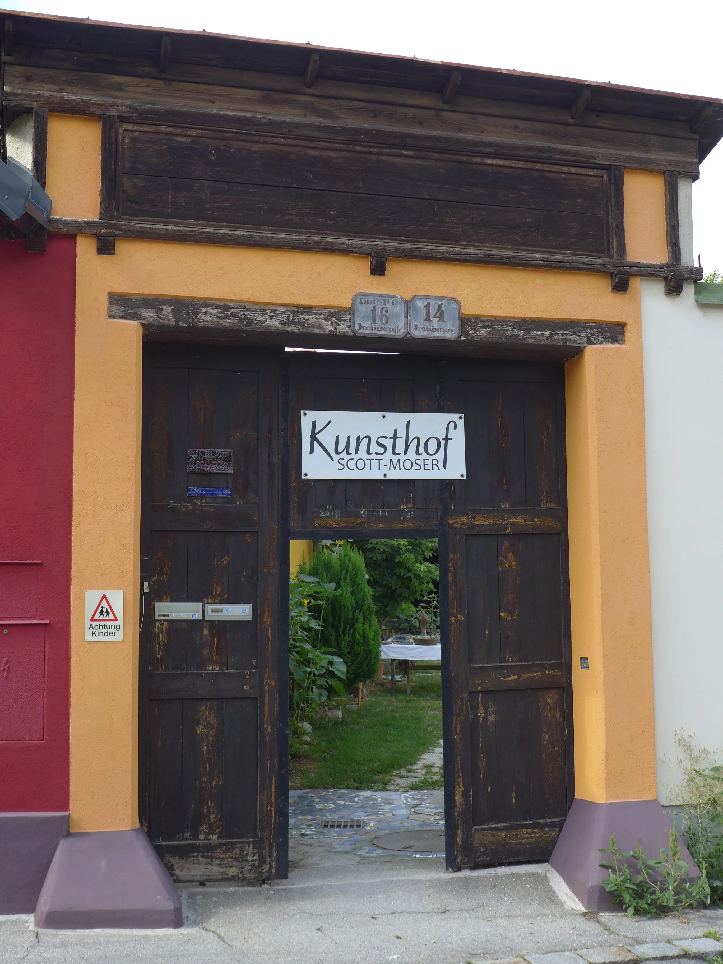 Entrance gate with dark wooden doors labeled Kunsthof Scott-Moser, opening to a garden with greenery and a table inside.