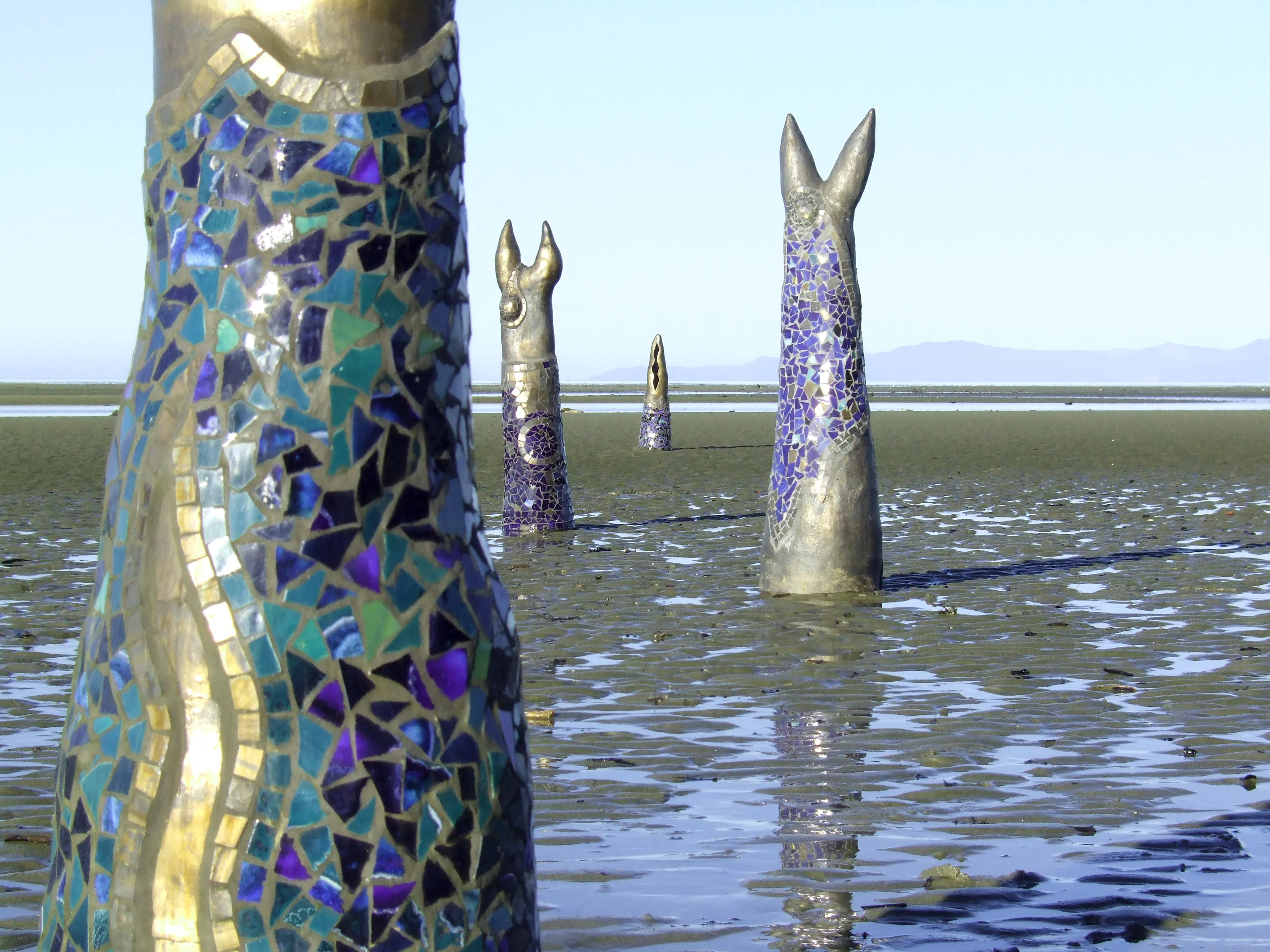 Four large, decorated animal head sculptures partially submerged in shallow water and mudflats with distant mountains under a clear sky.
