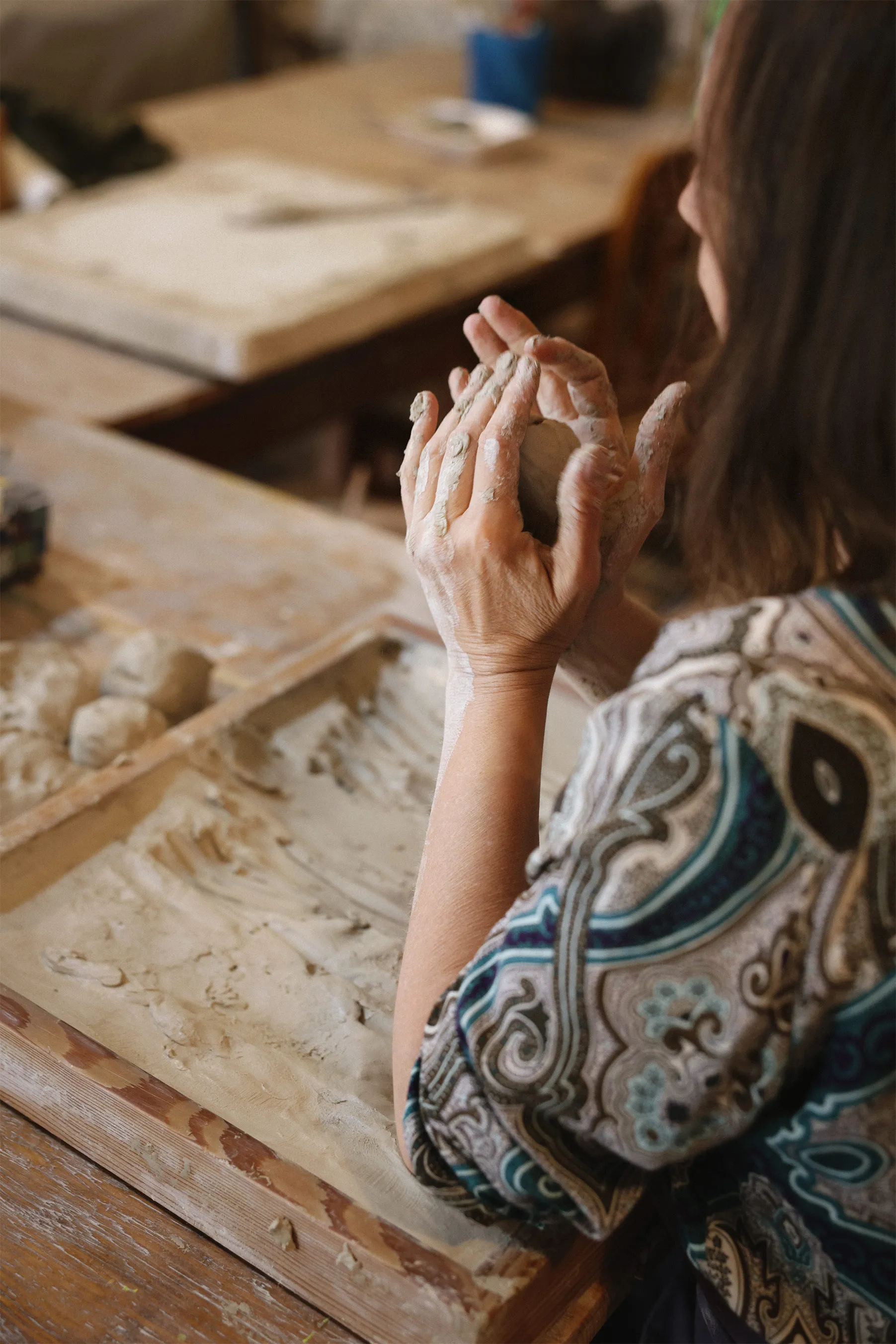 Person shaping a ball of clay with hands covered in clay at a wooden table with pottery tools.