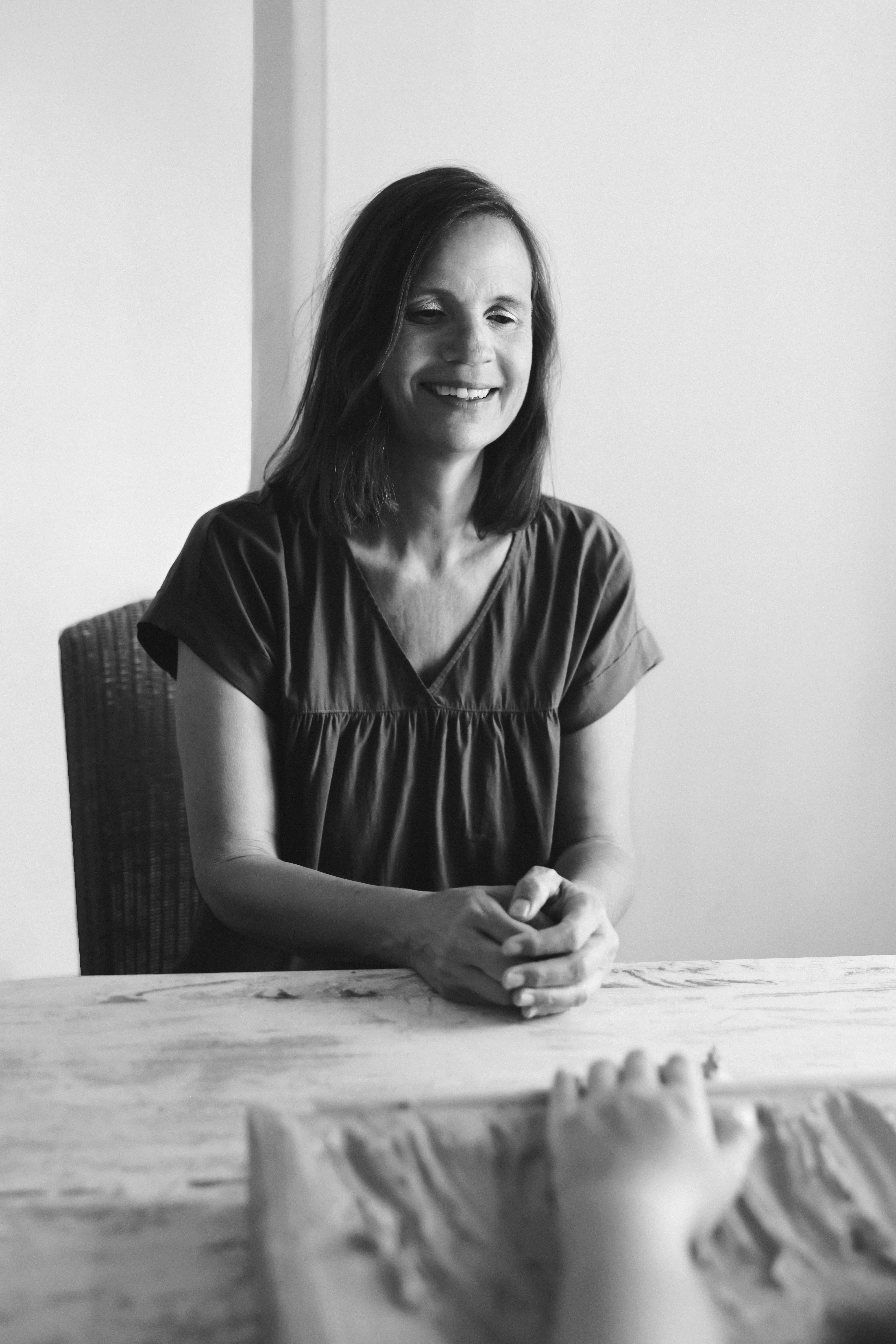 Smiling woman sitting at a table with hands clasped, looking at a child’s hand on a textured surface in foreground.