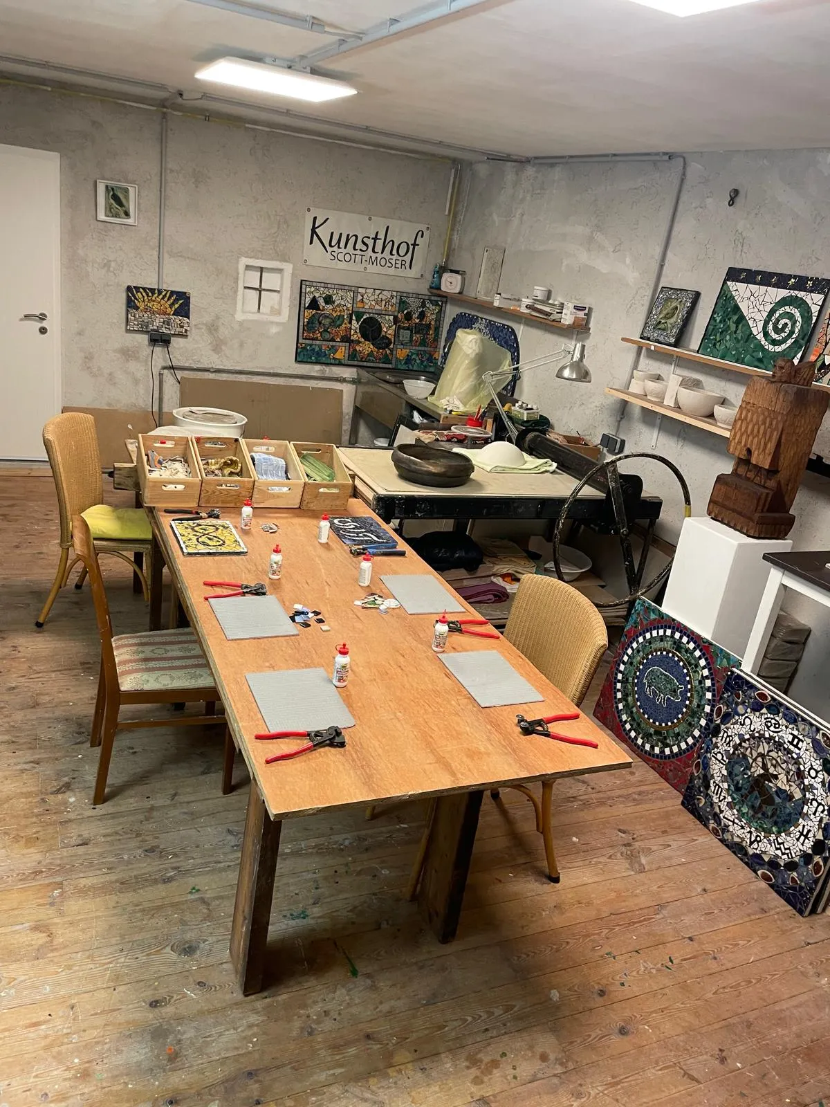 Art studio with a wooden table arranged for mosaic crafting with tools, glue bottles, and materials, surrounded by mosaic artworks and rustic decor.