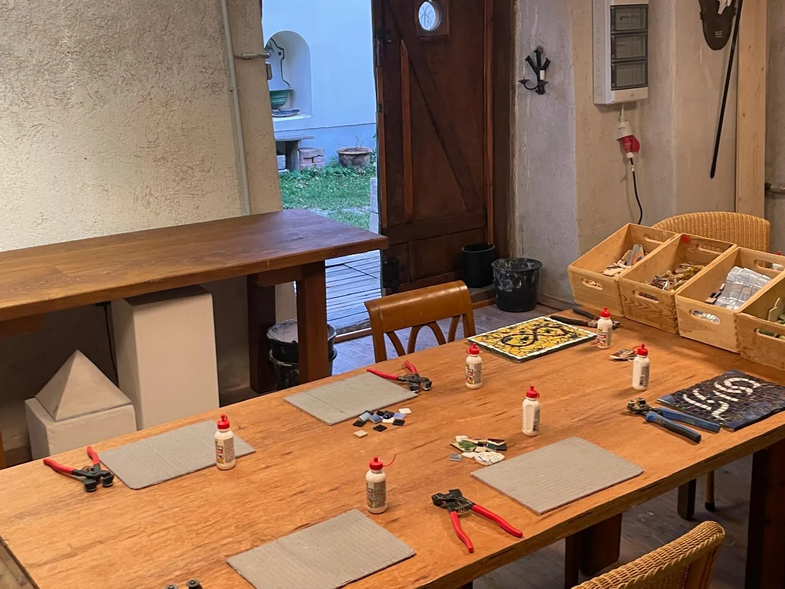 Wooden table set for mosaic crafting with glue bottles, tile cutters, small tile pieces, and woven chairs in a workshop room.