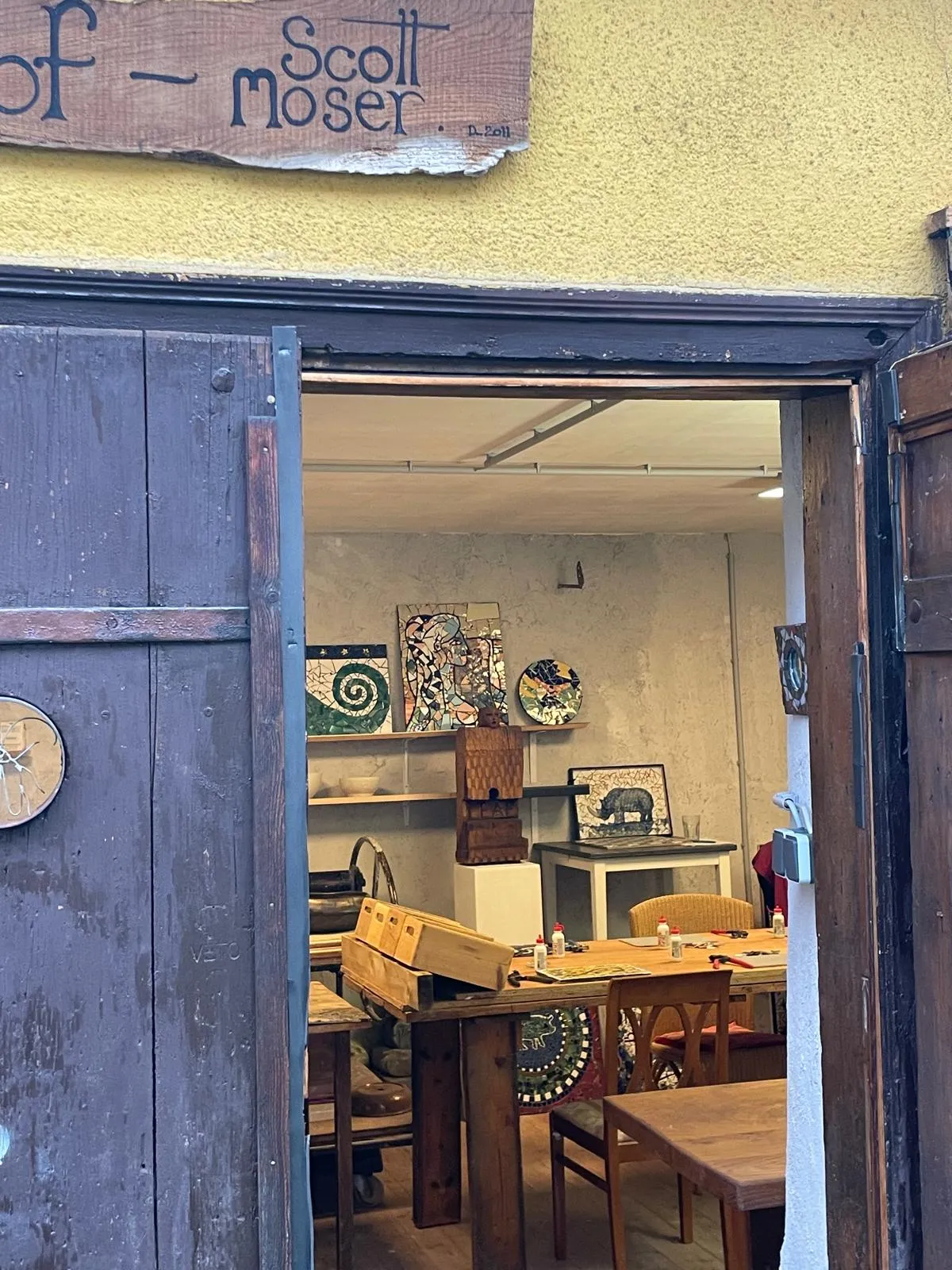 View through an open wooden door into an art studio with mosaic artworks, wooden tables, chairs, and crafting supplies.