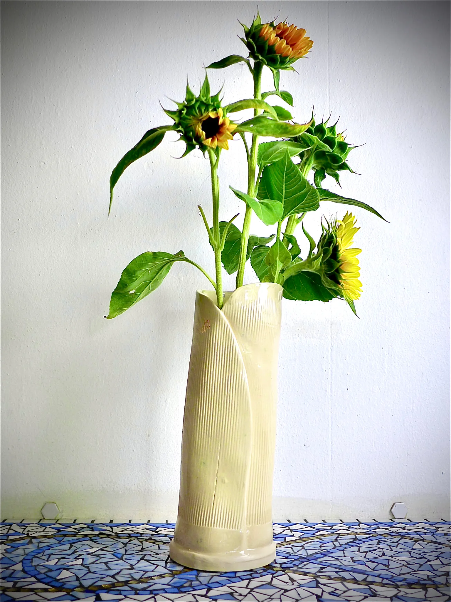 Cream-colored textured vase holding five sunflower stems with green leaves against a plain white wall on a blue and white mosaic surface.