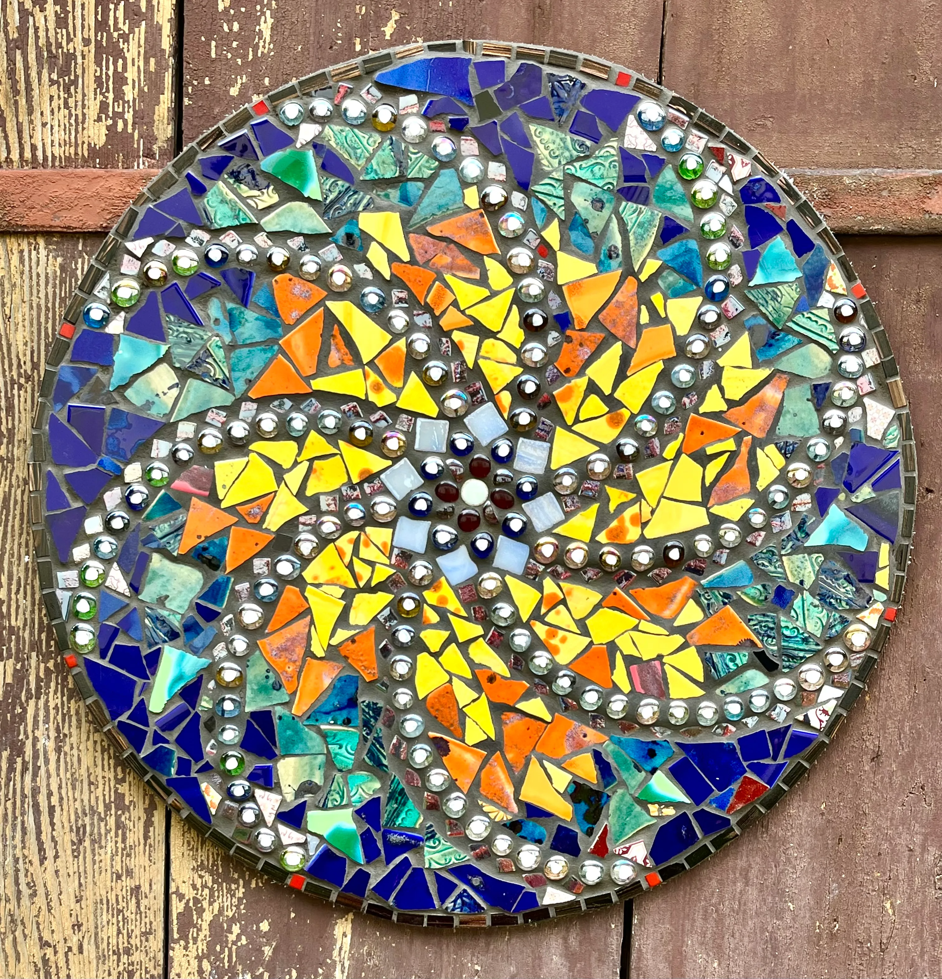 Round mosaic artwork with colorful triangular and irregular tiles in yellow, orange, blue, and green, decorated with small mirrored circular pieces, mounted on weathered wooden boards.