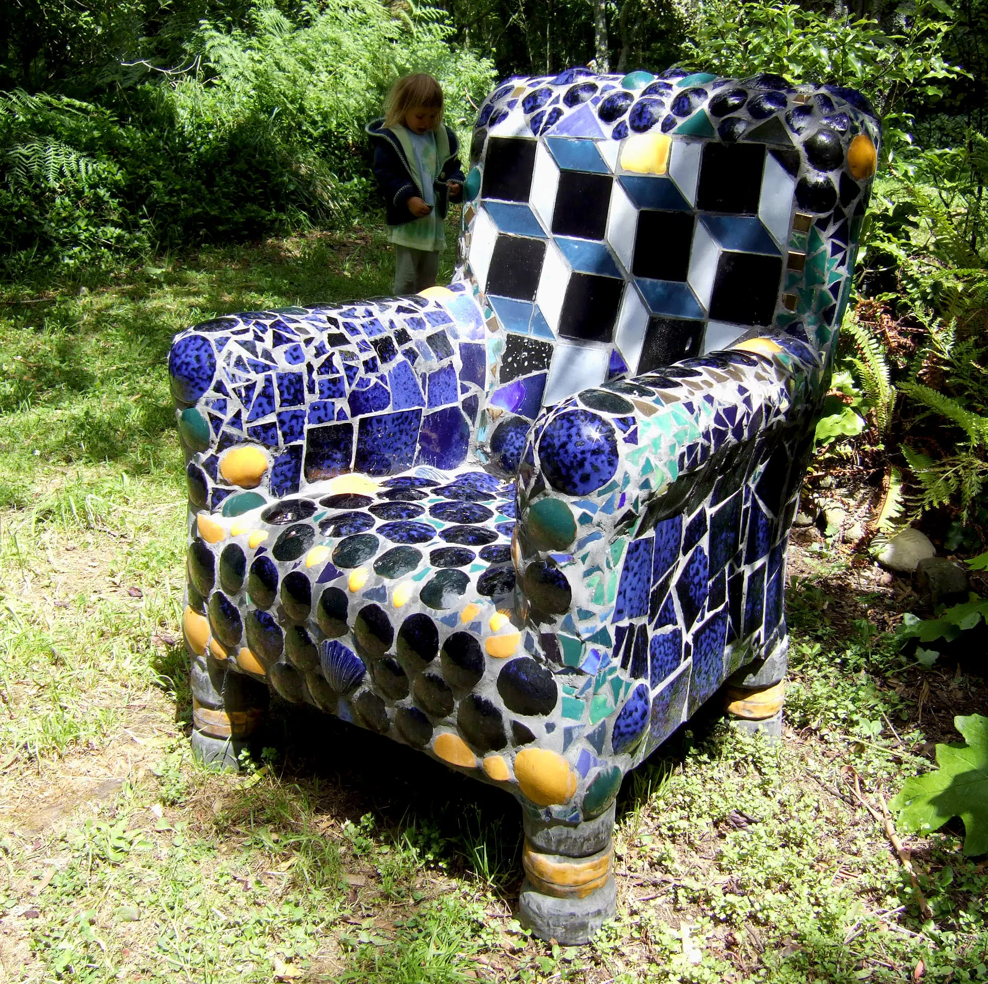 Colorful mosaic chair with geometric patterns and circular tiles placed outdoors on grass, with a child in the background.