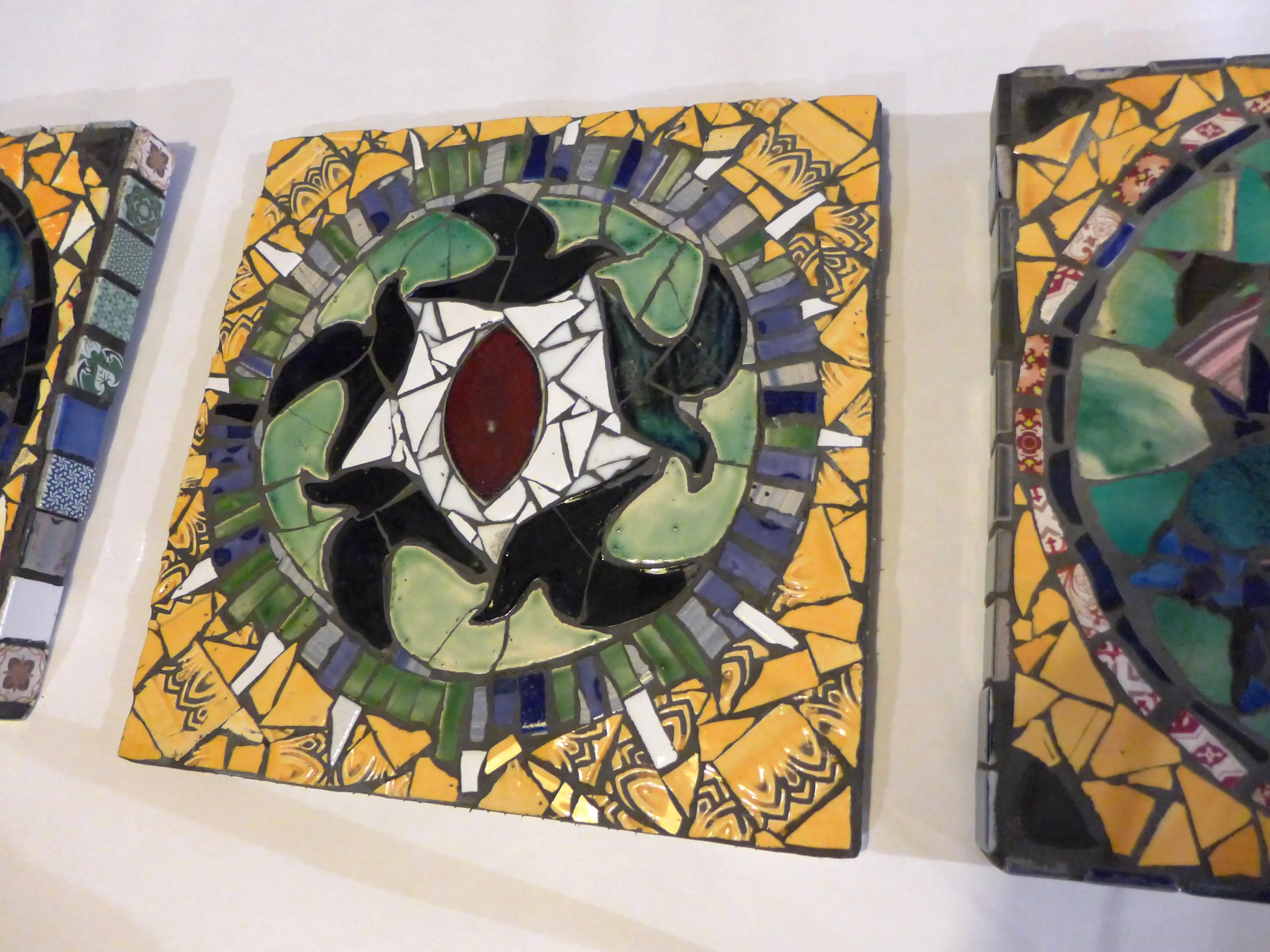 Square mosaic tile with black, green, white, and red shapes forming a circular pattern surrounded by yellow and blue bordered fragments.