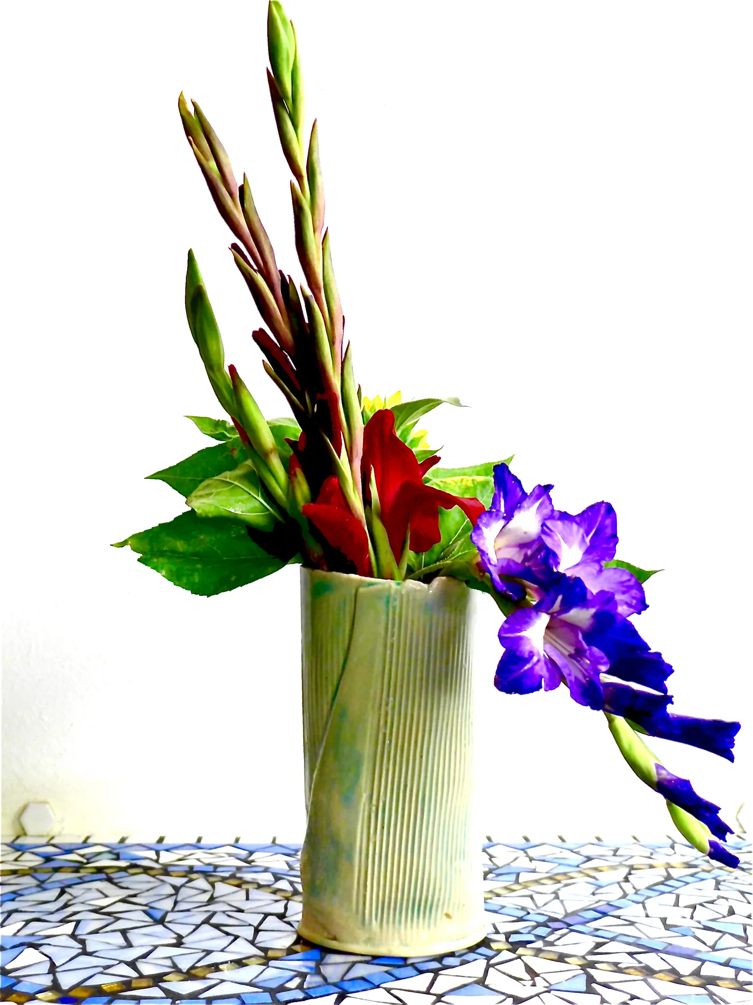 Ceramic vase with vertical ridges holding red, purple, and green flowers on a blue and white mosaic table.