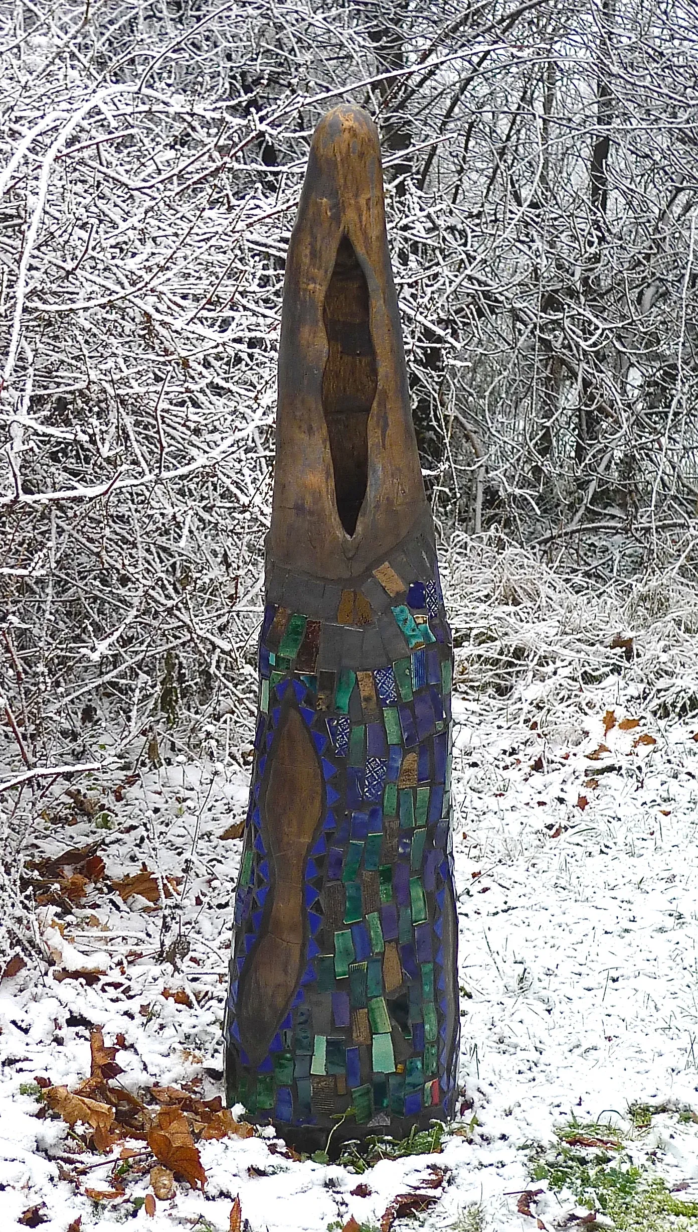 Tall wooden sculpture with mosaic tiles in green and blue standing on snow-covered ground with leafless, snow-dusted bushes in the background.