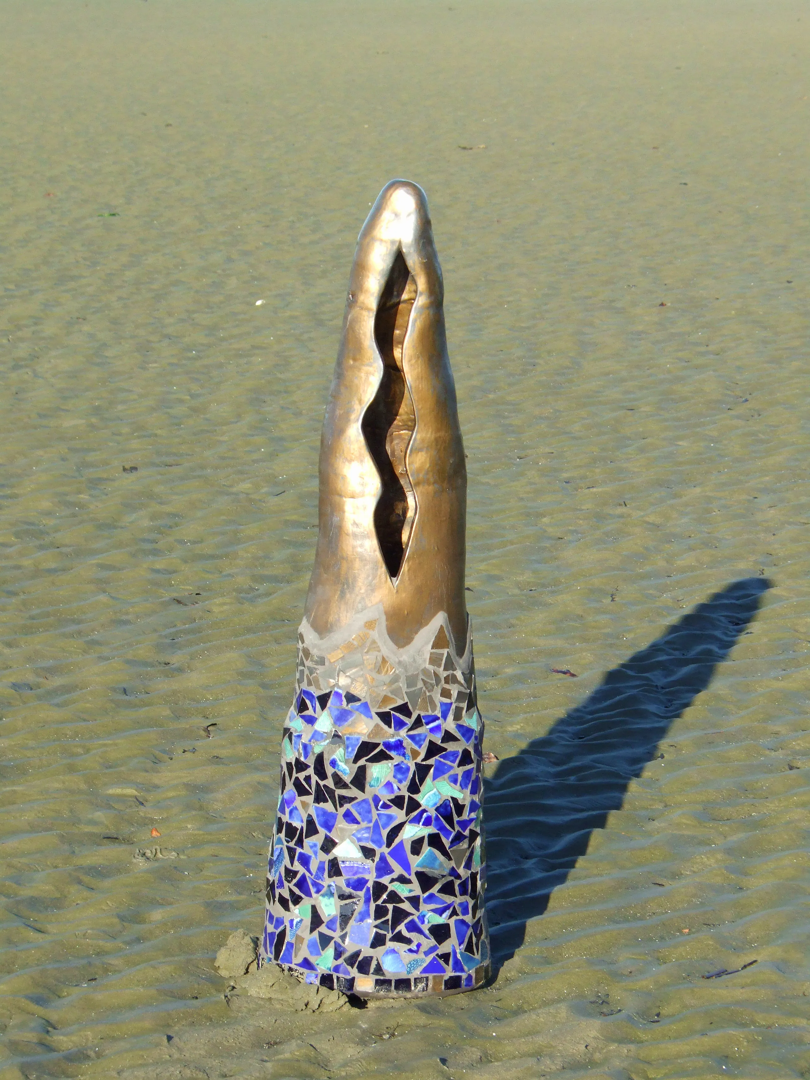 Tall sculpture on sandy ground with a mosaic base and an open, curved top casting a shadow.