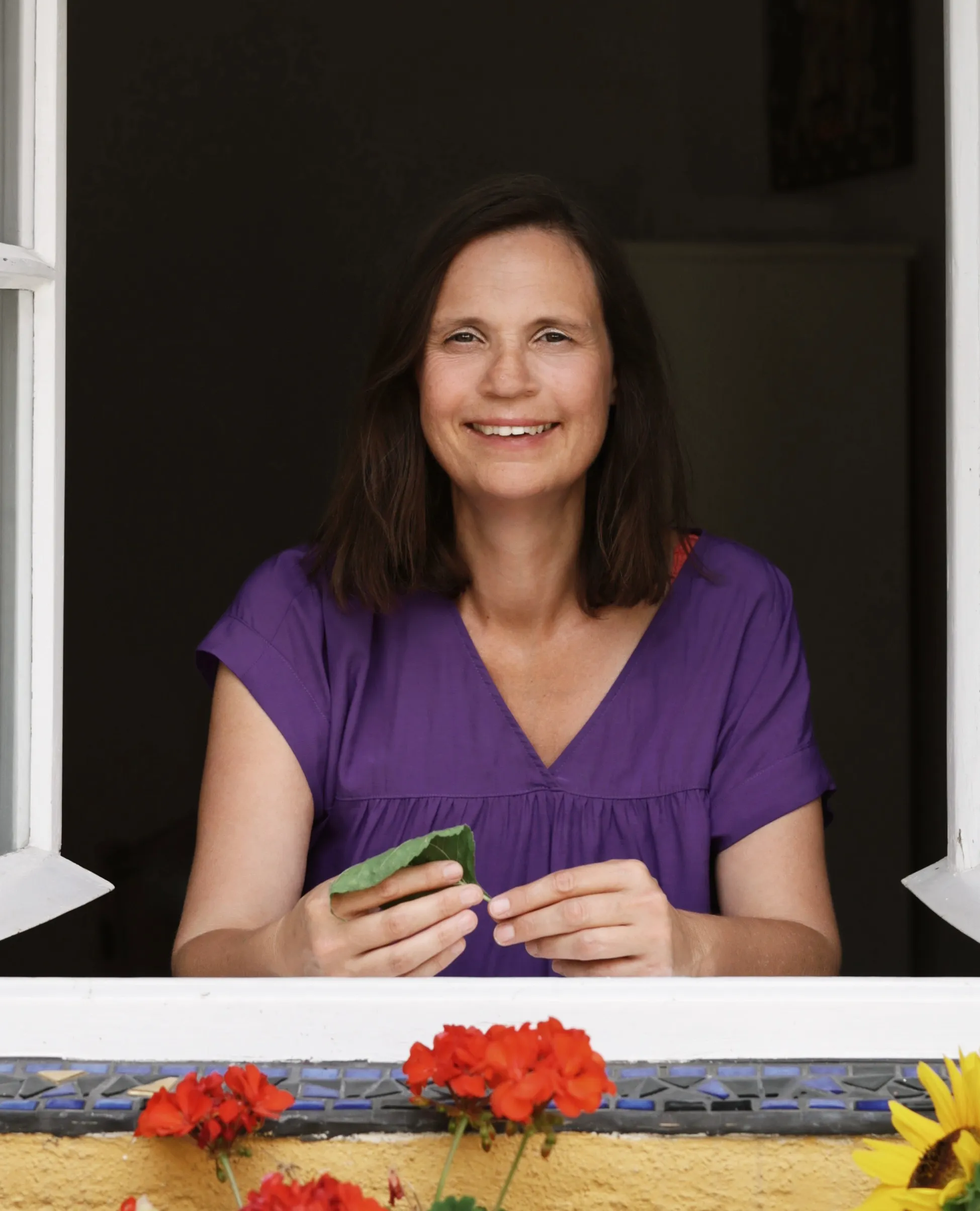 Lächelnde Frau mit braunem Haar in lila Bluse sitzt an offenem Fenster und hält grünes Blatt.