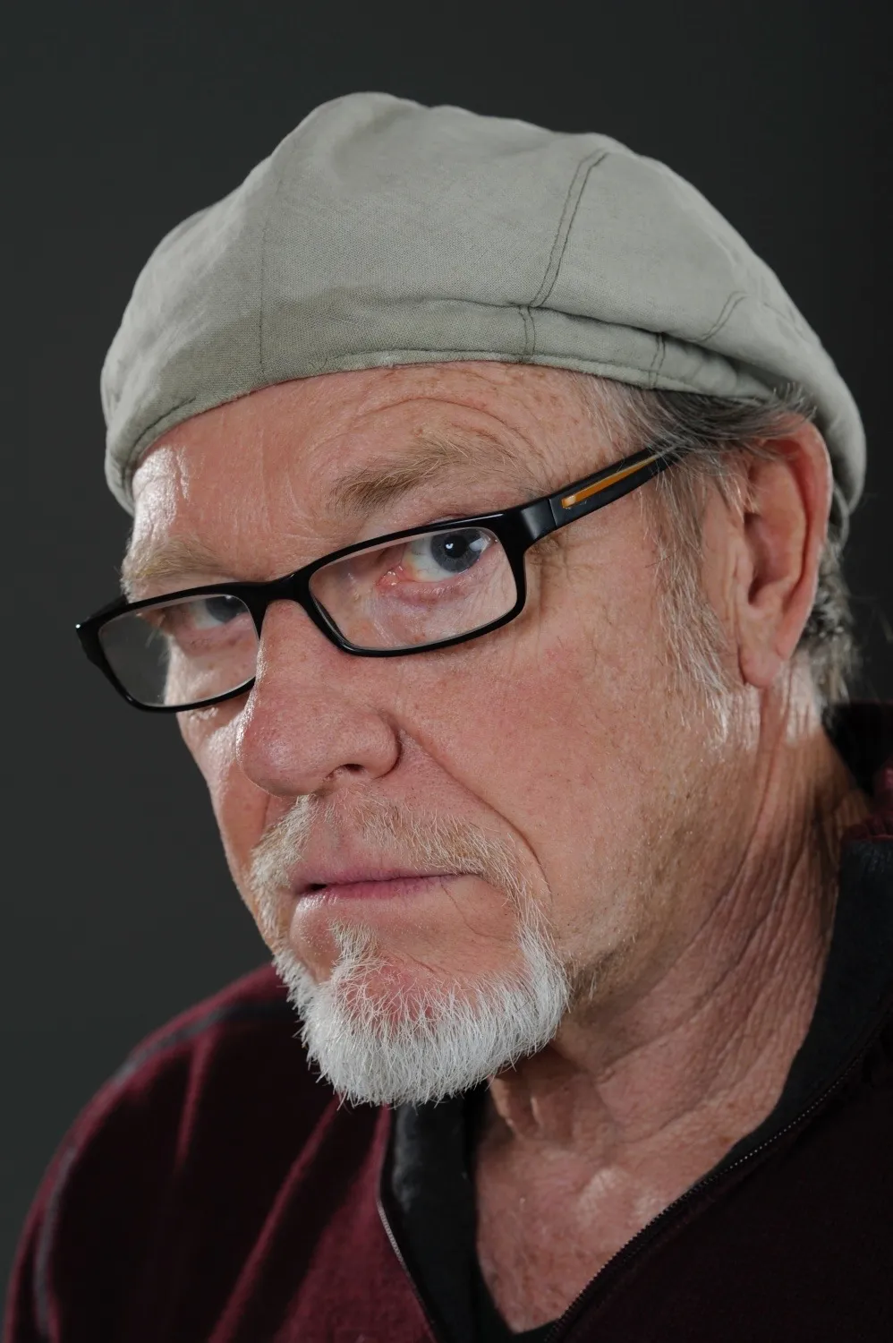 Porträt eines älteren Mannes mit grauem Bart, schwarzen Brillengestell und grauer Mütze vor dunklem Hintergrund.