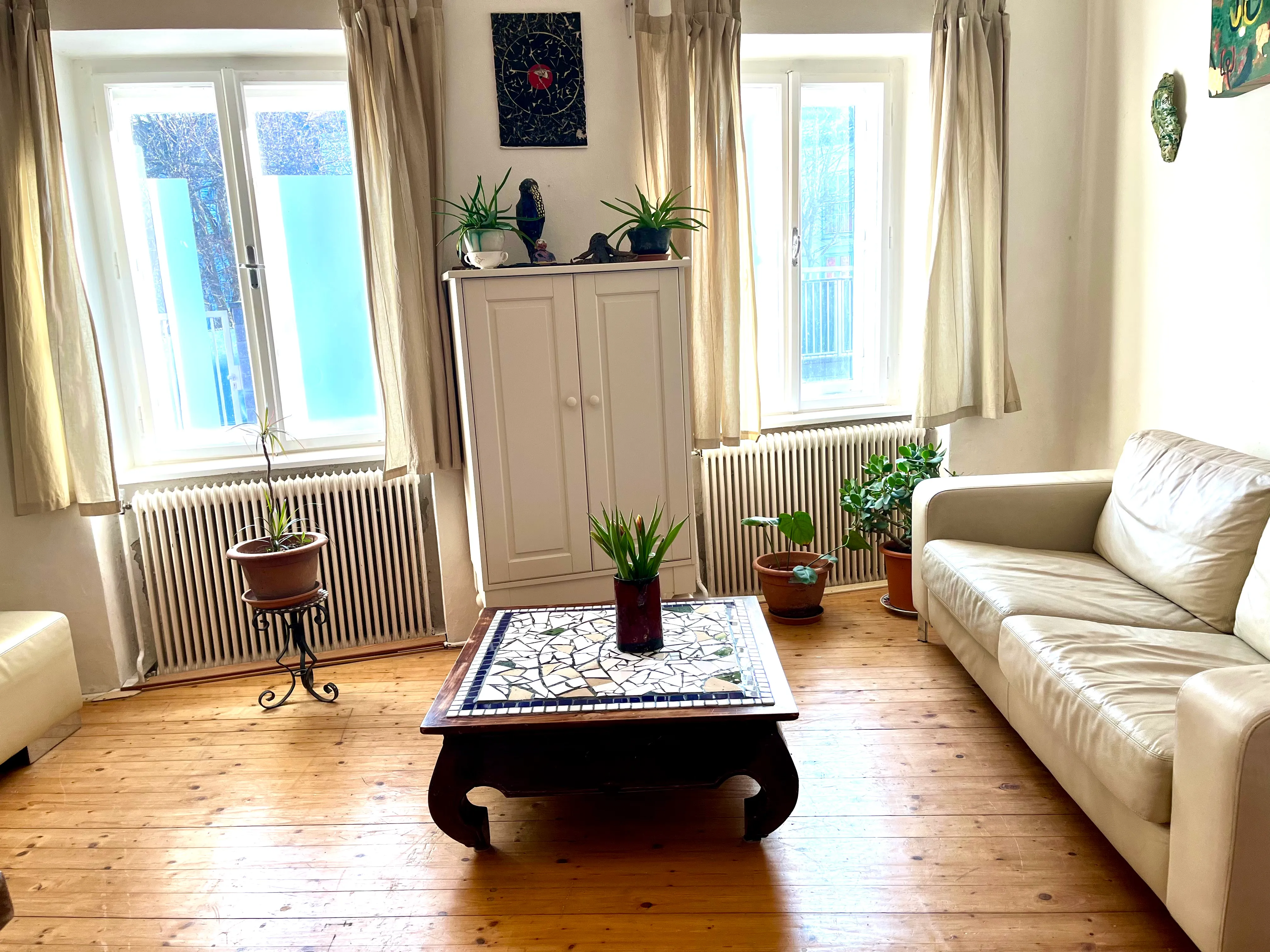 Helles Wohnzimmer mit hellem Ledersofa, einem niedrigen Holztisch mit Mosaikplatte und mehreren Pflanzen vor Fenstern mit hellen Vorhängen.