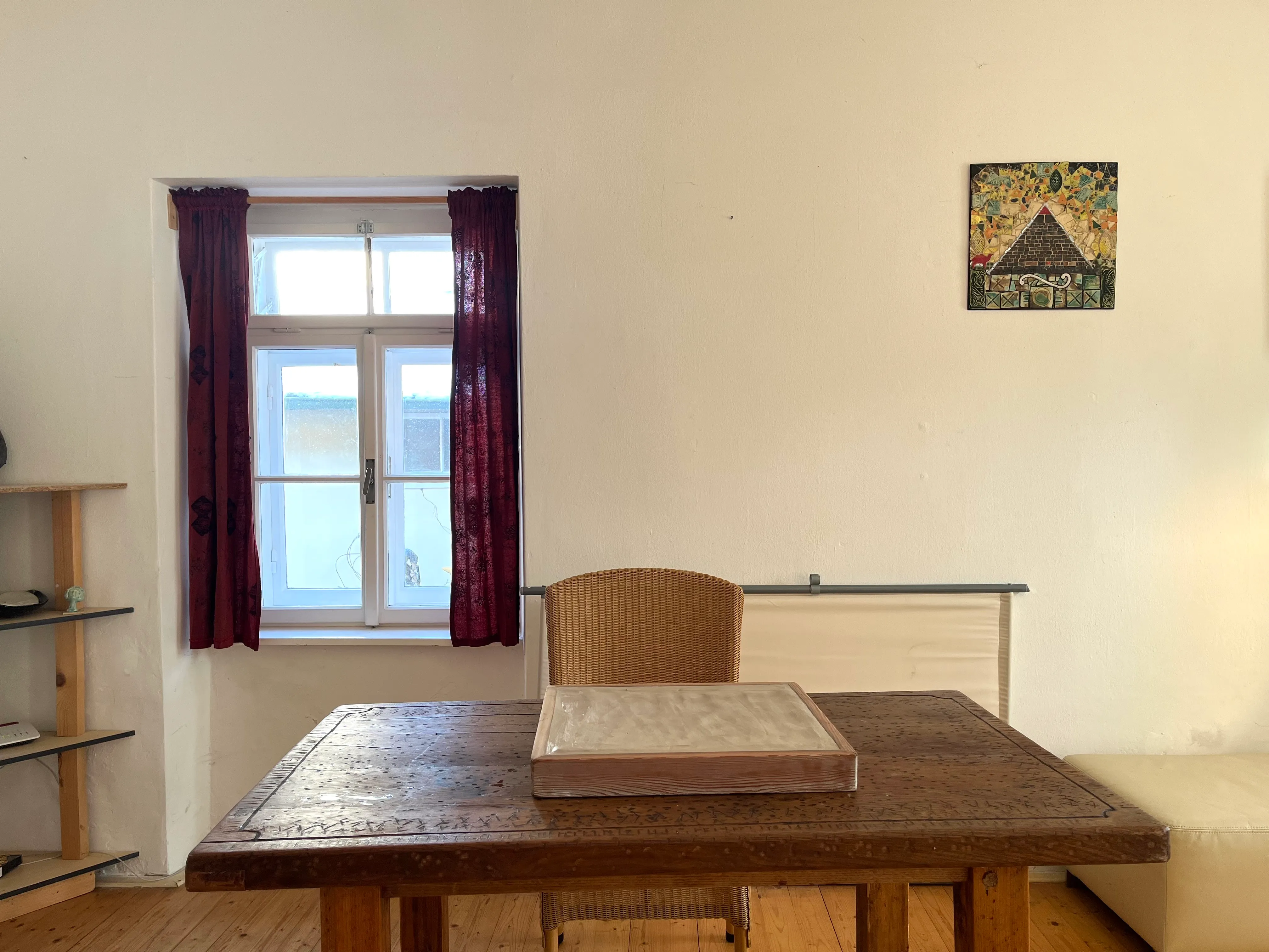 Ein helles Zimmer mit einem Holzschreibtisch, einem Korbstuhl, einem Fenster mit roten Vorhängen und einem kleinen Bild an der Wand.