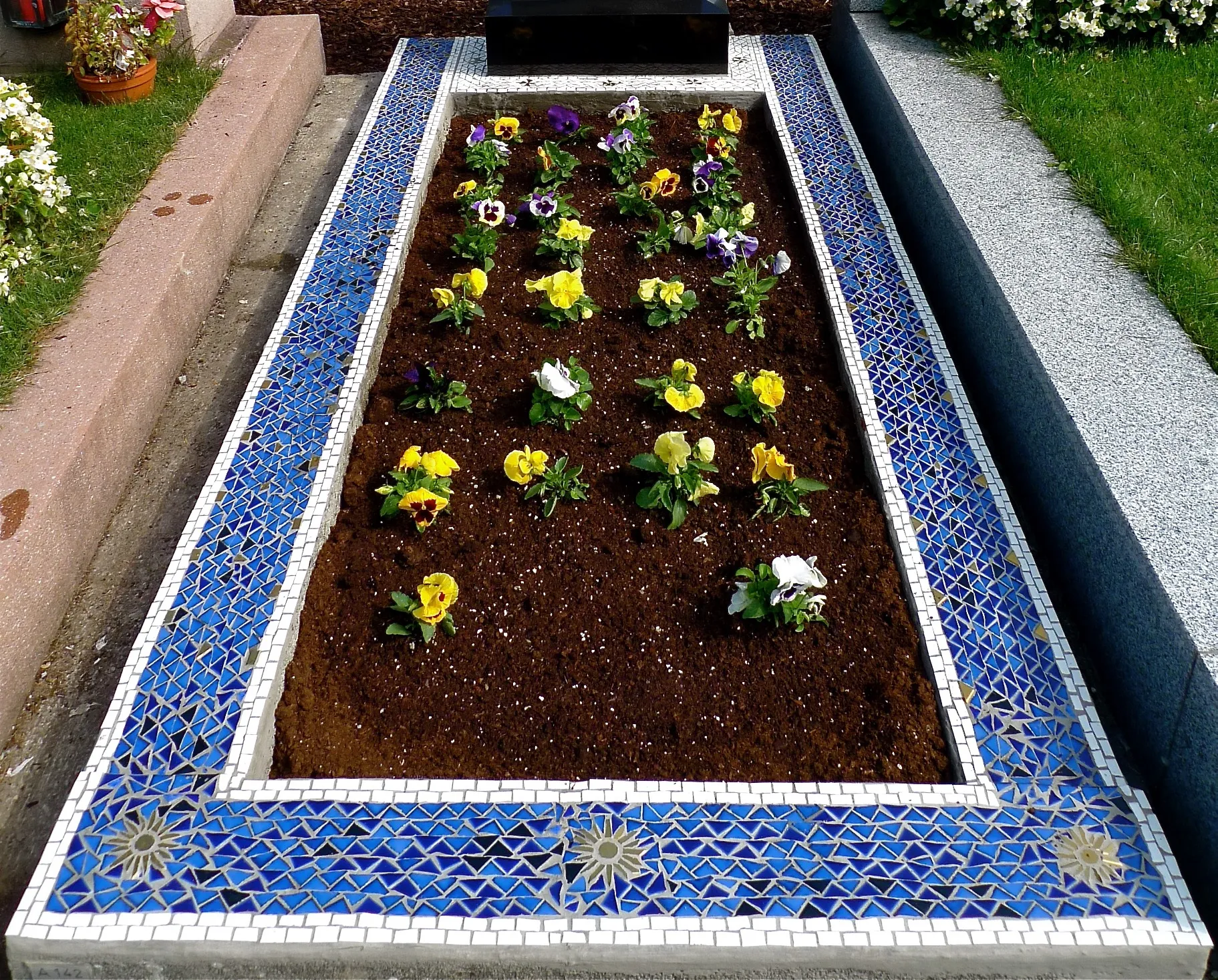 Grab mit Blumendeckung aus frisch gepflanzten gelben, weißen und violetten Stiefmütterchen, umgeben von einem Rand aus blau-weißen Mosaikfliesen mit Sonnenmotiven.