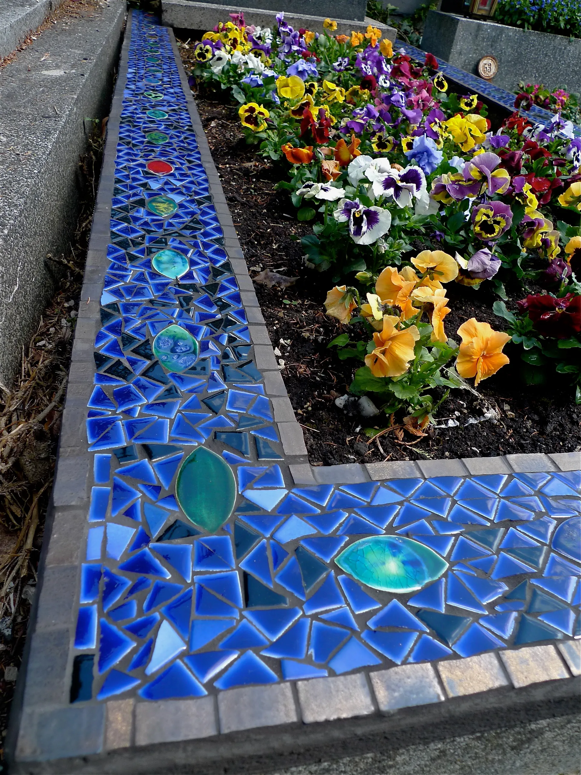 Grabumrandung mit einem blauen Mosaikmuster aus Keramikfliesen und bunten Stiefmütterchen im Blumenbeet.