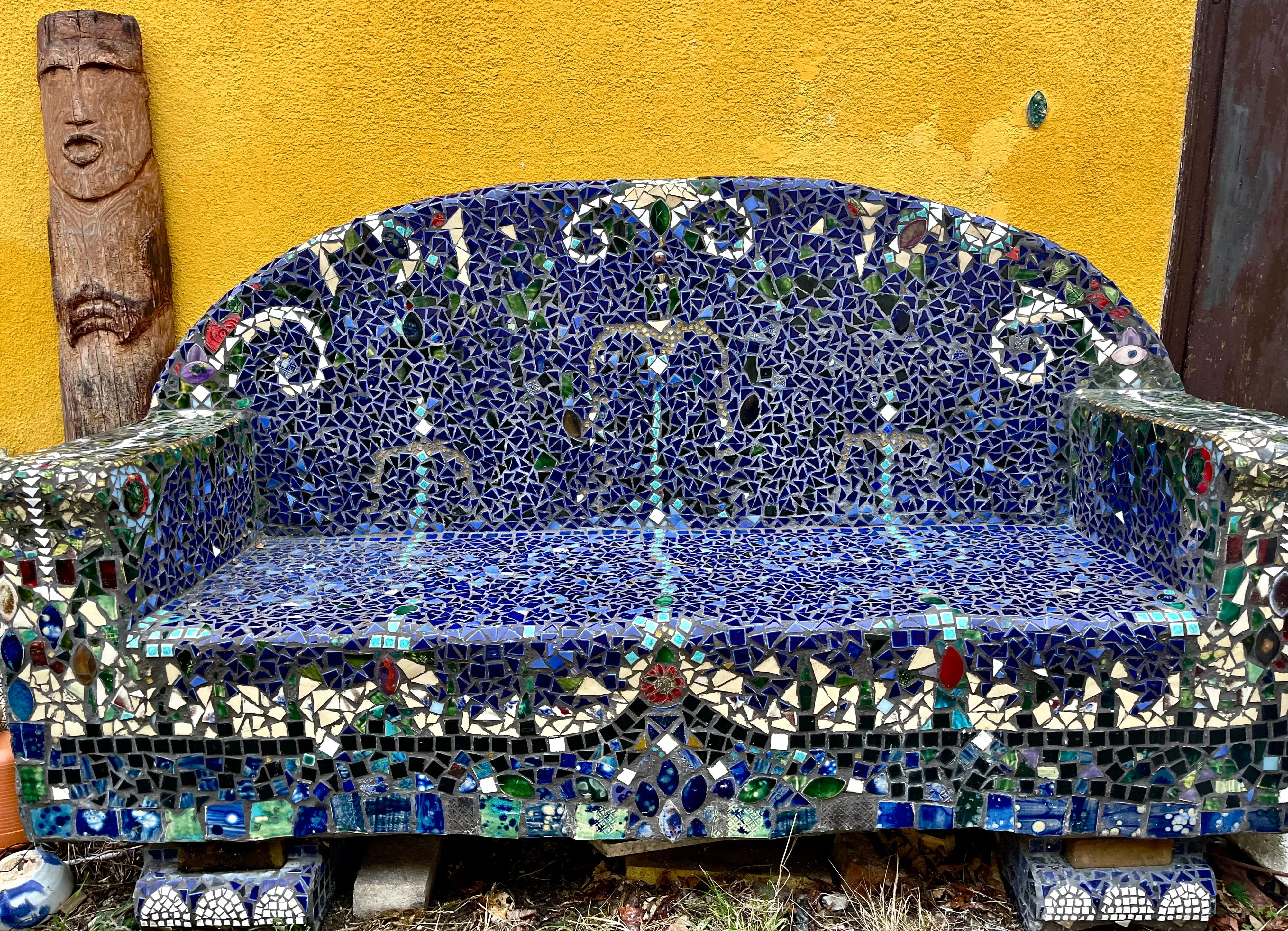 Bank aus blauen und bunten Mosaikfliesen vor gelber Wand mit geschnitzter Holzfigur links.