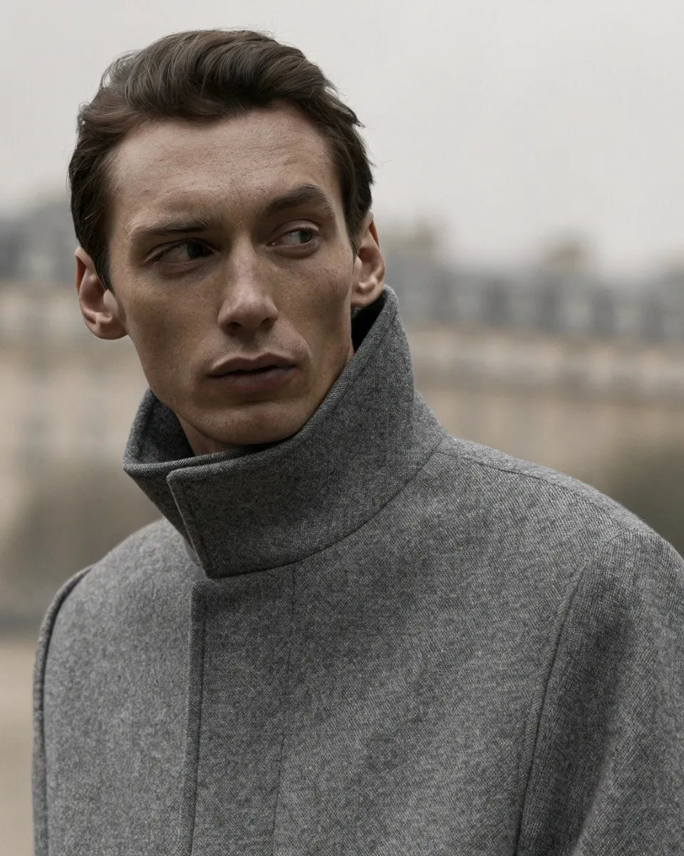 Contemplative man wearing a gray coat with the collar turned up, looking to his left against a blurred urban background.