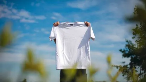 A person holding up a white t - shirt in front of a blue sky.
