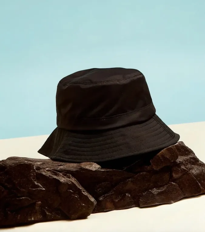 Black bucket hat resting on dark textured rocks against a light blue and white background.