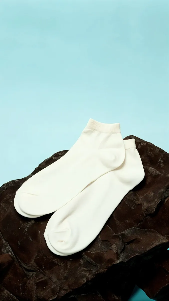Pair of minimalist white ankle socks placed on a textured dark brown fabric against a light blue background.