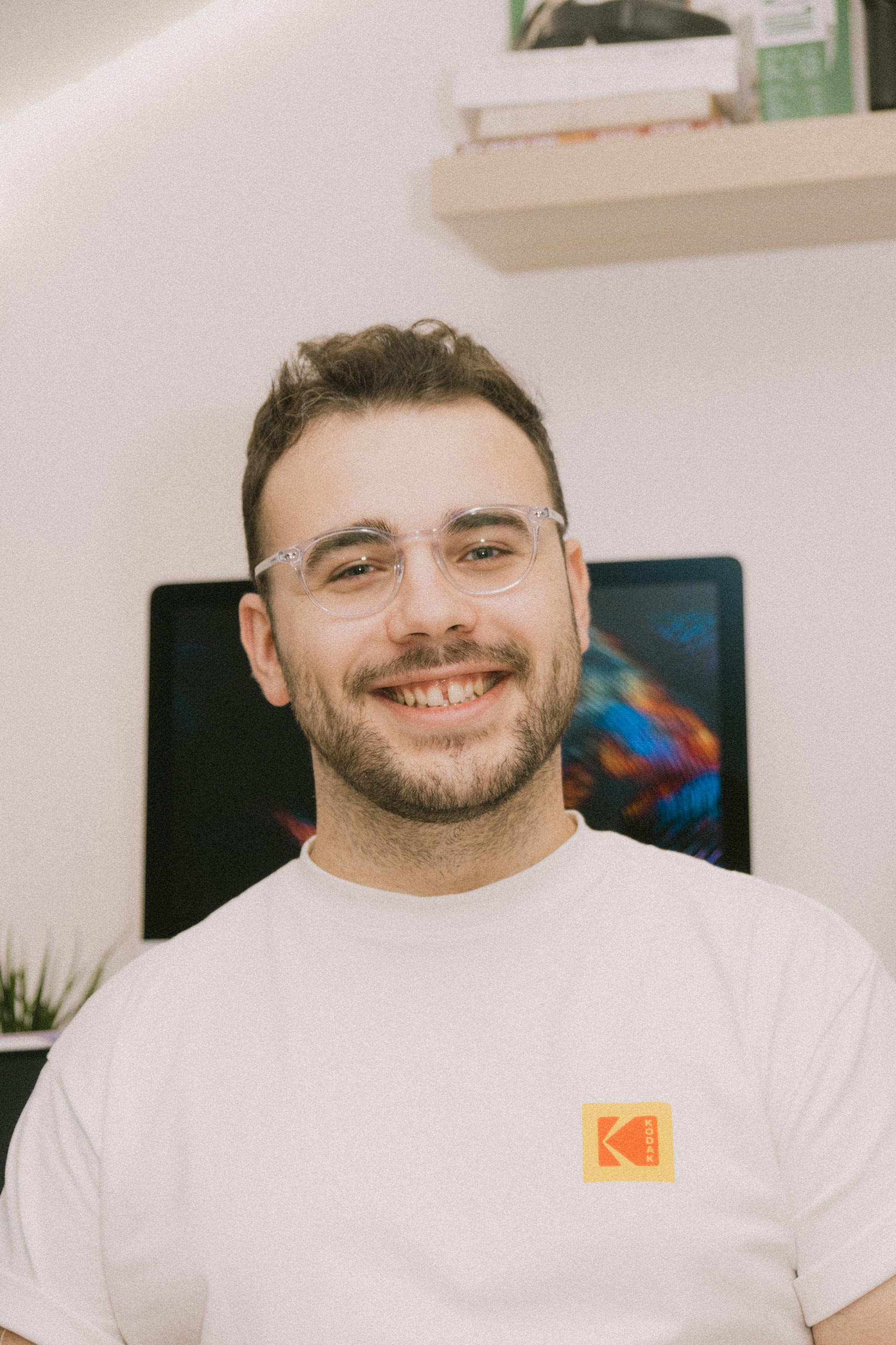 Smiling man with short curly hair and glasses wearing a white Kodak t-shirt in a home office.